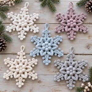 May include: Five crocheted snowflake ornaments in white, light blue, dusty pink, and gray. Each six-pointed star features a central cluster of bobble stitches surrounded by intricate, textured loops and a small hanging loop at the top.