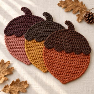 May include: Three acorn-shaped crochet coasters featuring dark brown caps and bodies in shades of muted pink, mustard yellow, and burnt orange. Each coaster has a small stem at the top and a scalloped edge where the cap meets the nut body.