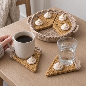Puede incluir: Un juego de posavasos de ganchillo con forma de porciones de tarta, cada uno con un borde de corteza dorada y un pequeño copete de crema blanca. El set incluye un soporte redondo con forma de tarta y dos posavasos triangulares.