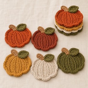 May include: A collection of six crocheted pumpkin-shaped coasters in various colors including orange, mustard yellow, cream, and olive green. Each coaster features a small brown stem and a single green leaf attached to the top of the pumpkin shape.