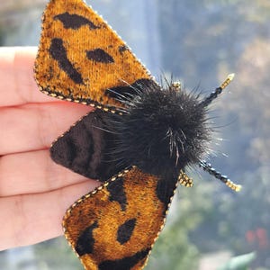 May include: A moth brooch featuring velvet wings in a golden-brown and black leopard-like pattern, edged with gold beads. The central body is composed of fluffy black fur, and the antennae are crafted from black and gold beads on thin wires.