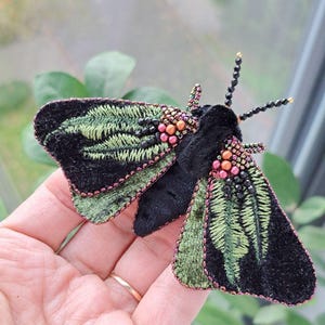 Peut inclure: Une broche en forme de papillon de nuit avec des ailes en velours noir ornées de motifs de fougères brodés en vert, rehaussées de perles métalliques cuivrées et noires. Le corps est en velours noir avec deux antennes terminées par des perles dorées.
