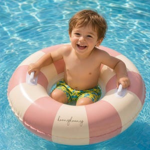 May include: An inflatable circular pool float featuring alternating dusty pink and cream-coloured segments. The float includes two white plastic handles and the text koongkoongs printed in a cursive black font on one of the cream-coloured sections.
