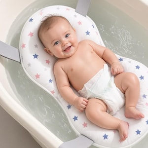 Puede incluir: Un cojín de baño para bebé de color blanco con un patrón de pequeñas estrellas azules y rosas. El cojín tiene forma para sostener a un infante e incluye correas grises para sujetarlo a una bañera, proporcionando una superficie suave.
