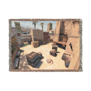 May include: A woven throw blanket featuring a detailed depiction of a desert-themed video game map. The scene includes tan stone buildings, palm trees, wooden crates scattered across a sandy courtyard, and a red letter A symbol on the ground.