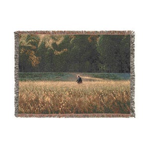 May include: A rectangular woven throw blanket featuring a landscape scene of a person walking through a golden field at sunset. The image displays a dark forest line in the background, textured fringe edges, and a detailed tapestry-style weave.