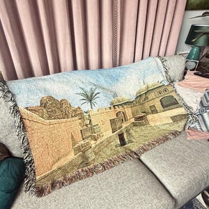 May include: A rectangular woven throw blanket featuring a detailed landscape scene of a desert fortress with tan stone walls, a single palm tree, and domed structures under a light blue sky. The blanket has a decorative fringe along the edges.