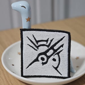 May include: A square embroidered patch featuring a white background with a black stitched graphic design. The patch has a thick black border and is displayed resting against a light blue ceramic figure shaped like a long-necked creature.