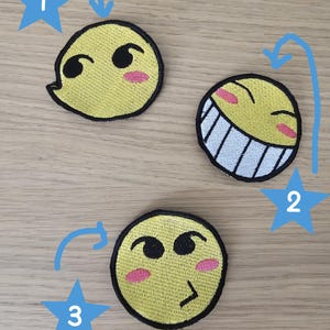 May include: Three circular embroidered patches featuring yellow faces with black outlines. Patch 1 shows a side-glancing expression. Patch 2 displays a wide, toothy grin with closed eyes. Patch 3 depicts a face with a smirk and pink cheek accents.