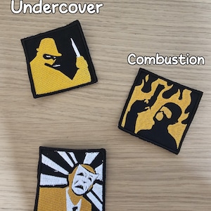 以下が含まれることがあります： 黒い縁取りの正方形の刺繍ワッペン3枚。左上のワッペンは帽子をかぶった黄色い人物がナイフを持っている図で「Undercover」と書かれています。右上のワッペンは黄色い炎を背景にした黒いシルエットで「Combustion」と書かれています。下のワッペンは仮面の図で「Costume Contest」と書かれています。