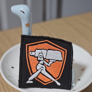 May include: A square embroidered patch featuring a black background with an orange shield outline. Inside the shield is a white graphic of a tripod-mounted camera or sensor device. The patch is displayed on a light blue ceramic dinosaur figurine.