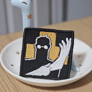 May include: A square embroidered patch featuring a black silhouette of a figure wearing round glasses against a gold background. A white arm and hand are extended across the foreground. The patch has a thick black border and is stitched in detail.