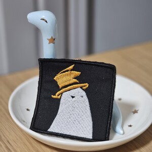 May include: A square black embroidered patch featuring a white ghost wearing a gold top hat. The patch is resting against a light blue ceramic figure of a long-necked creature, which is placed inside a white dish decorated with small gold stars.