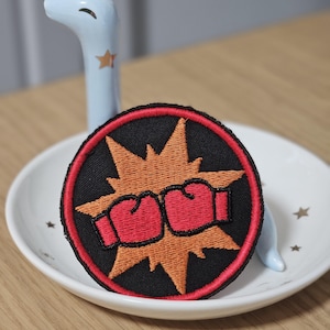Op de afbeelding: Een ronde geborduurde patch met een zwarte achtergrond en een rode rand. Het ontwerp toont twee rode bokshandschoenen die in het midden botsen, geplaatst tegen een grillige, oranje ster die een dynamisch stripboekachtig impacteffect creëert.
