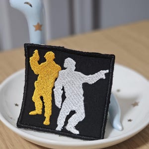May include: A square black embroidered patch featuring two figures in a dancing pose. One figure is stitched in bright yellow thread, while the other is stitched in white thread. The patch has a black border and a flat, rectangular shape.