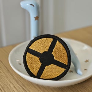 May include: A circular embroidered patch featuring a black border and a central black cross design that divides the patch into four golden yellow segments. The patch is positioned in front of a light blue ceramic figurine with gold star accents.