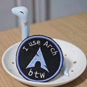 May include: A circular embroidered patch with a black background and a light blue border. The patch features a light blue stylized letter A logo in the center with the white embroidered text I use Arch btw surrounding the logo in a circular pattern.