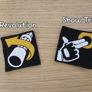 May include: Two square black embroidered patches with white and yellow thread details. The left patch features a white tube with a yellow arrow and the text Revolution. The right patch shows a white gloved hand pointing at a yellow ring with the text Show Trial.
