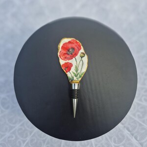 Op de afbeelding: Een flessenstop met een witte, onregelmatig gevormde bovenkant, versierd met een illustratie van felrode klaprozen en groen blad. De randen zijn afgewerkt in goud. De basis is gemaakt van zilverkleurig metaal met zwarte ringen.