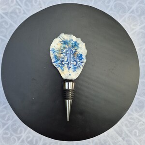 Op de afbeelding: Een flessenstop met een natuurlijke, onregelmatige witte schelp als bovenkant, versierd met een blauwe inktillustratie van een zeewezen. De stop heeft een taps toelopende, zilverkleurige metalen basis met zwarte rubberen ringen voor een goede pasvorm.