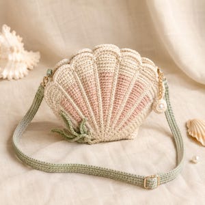 Peut inclure: Un sac bandoulière en crochet en forme de coquillage, couleur crème et rose pâle, doté d'une sangle vert sauge. Le sac présente un bord festonné, une petite décoration en forme d'algue verte et une tirette dorée avec une perle blanche.