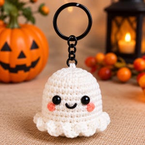 May include: A white crocheted ghost keychain featuring a rounded body with a ruffled bottom edge, two black plastic bead eyes, a stitched black mouth, and two small pink circular cheeks. The ghost is attached to a black metal split ring.