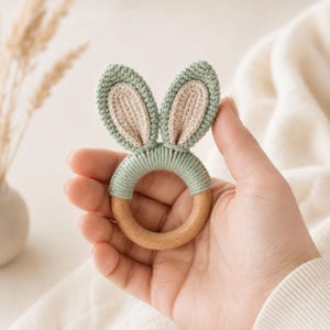 Op de afbeelding: Een rammelaar voor baby's met een gladde, ronde houten ring die gedeeltelijk is omwikkeld met saliegroen garen. Aan de bovenkant zitten twee rechtopstaande, gehaakte konijnenoren met saliegroene randen en lichtbeige, textuurrijke centra.