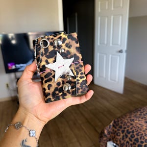 May include: A rectangular wallet featuring a brown and black leopard print pattern. The front is decorated with a white star-shaped patch, silver-toned metal studs, a thin pink elastic band, and a silver metal ring attached to a small ball chain.