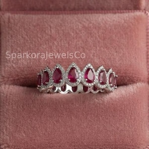 May include: A silver ring featuring a row of pear-shaped red rubies, each surrounded by a halo of small white diamonds. The ring is displayed inside a dusty pink velvet box with the text SparkoraJewelsCo visible on the inner lining.