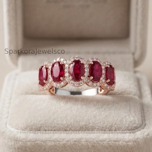 Può includere: Un anello in oro rosa con cinque rubini rossi taglio ovale disposti in fila, ciascuno circondato da un alone di piccoli diamanti bianchi. L'anello è centrato in un portagioie in velluto beige con il testo SparkoraJewelsco visibile a sinistra.