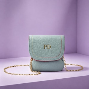 May include: A light blue pebbled leather crossbody bag with a fold-over flap featuring the gold embossed initials PD. The bag includes a thin gold metal chain strap and visible red stitching along the edges of the flap and main body.