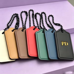Può includere: Un set di sette etichette per bagagli rettangolari nei colori crema, beige, marrone, rosso, verde, blu e nero. Ogni etichetta presenta una catenella a palline nera abbinata e un bordo in rilievo. L'etichetta nera reca le lettere dorate PD in rilievo.