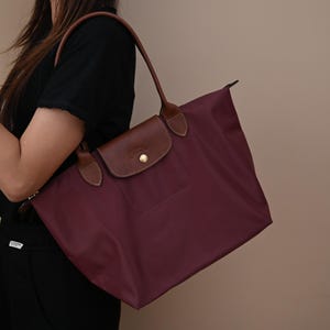 Puede incluir: Un bolso tote de color burdeos intenso con cuerpo de nailon liso y adornos de cuero marrón. El bolso incluye una solapa con cierre de botón a presión dorado, dos correas largas de cuero para el hombro y un cierre de cremallera superior.
