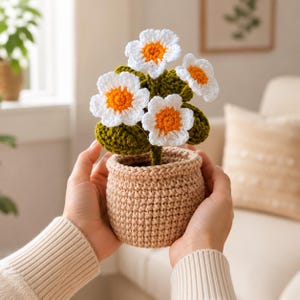 May include: A crocheted potted plant featuring three white flowers with bright orange centres and dark green leaves. The plant is housed in a textured, light tan crocheted pot with a thick, rounded rim, showcasing intricate yarn stitching.
