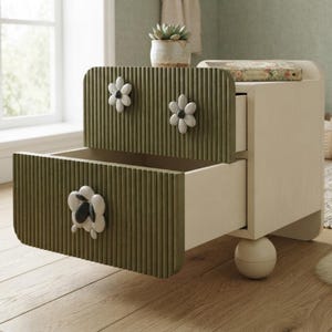 May include: A two-drawer nightstand with a light cream frame and olive green ribbed drawer fronts. The drawers feature white flower-shaped knobs with dark centers. The unit is supported by a single spherical leg and has a small potted plant on top.