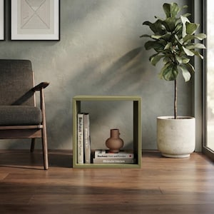 May include: A square olive green open shelf unit containing two books titled Bauhaus: Form and Function and Minimalist Interiors, along with a small brown ceramic vase. The shelf is positioned on a wooden floor next to a dark gray armchair.