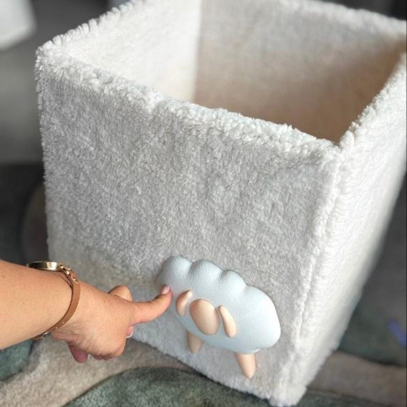 May include: A square storage bin made of white, textured, fleece-like fabric. The exterior features a three-dimensional, light blue, cloud-shaped applique with smooth, rounded edges and small, pale pink accents at the bottom.