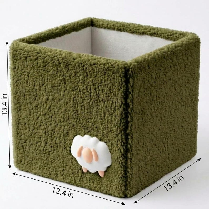 May include: A square storage bin covered in olive green textured fleece fabric. The exterior features a small white sheep applique with peach-colored accents. The interior is lined with a smooth off-white fabric. The bin measures 13.4 inches.