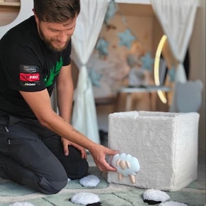 May include: A white, square-shaped storage bin made of textured, plush fabric. The front of the bin features a light blue, cloud-shaped applique with a small, beige sheep figure attached to the center. The bin has a soft, fuzzy exterior.