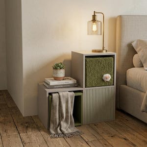 May include: A modular light gray storage unit with two open compartments and one closed compartment featuring a vertical ribbed door. The unit holds a green textured storage bin with a small white sheep accent, a succulent, and two books.