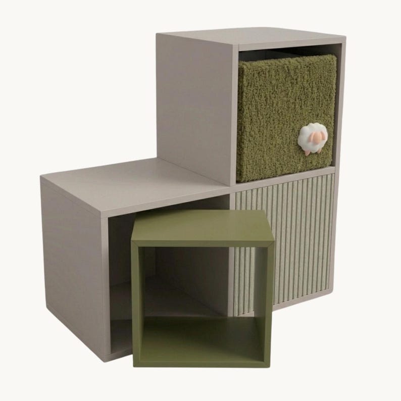 May include: A light gray modular storage unit with two open cube sections and two closed compartments. One compartment features a textured olive green fabric front with a small white sheep applique, while the other has a ribbed olive green panel.