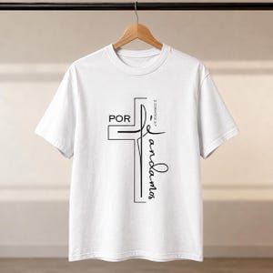 Peut inclure: Un t-shirt blanc à manches courtes avec un motif graphique noir représentant une croix. Le texte "POR" est imprimé horizontalement, tandis que "andamos" est écrit verticalement le long de la croix. "2 CORINTIOS 5:7" est visible.