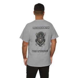 May include: A heather grey short-sleeve t-shirt featuring a graphic on the back. The design includes a central mechanical assembly of gears, a ball bearing, and wrenches, with the text PRECISION IN MOTION above and KEEP IT TURNING below.