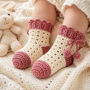 May include: A pair of cream and dusty rose crochet socks featuring a scalloped cuff, a lattice-patterned leg, and a solid toe section. The socks are accented with a decorative crochet bow and two small pom-poms on the side of the ankle.