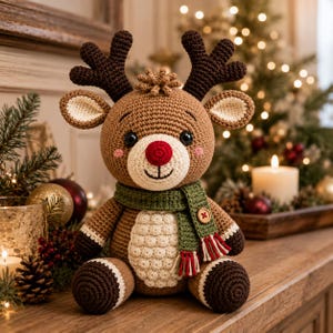 Puede incluir: Un peluche de reno tejido a ganchillo con cuerpo marrón claro, astas marrón oscuro y una nariz roja brillante. El reno lleva una bufanda verde con un botón de madera y flecos rojos y blancos, y tiene una barriga texturizada de color crema.