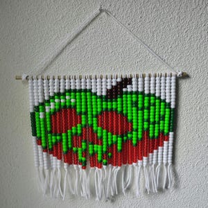 May include: A wall hanging made of white, green, and red plastic pony beads strung on white cord. The beads are arranged to form a graphic of a red apple with green highlights and a brown stem, suspended from a wooden dowel with a white string.