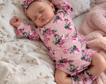 May include: A baby doll with closed eyes, wearing a long-sleeved pink bodysuit, matching socks, and a headband, all featuring a floral pattern with pink, white, and dark green flowers. The doll is positioned in a sleeping pose with arms raised.