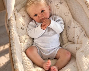 May include: A realistic reborn baby doll with blonde hair and blue eyes, wearing a long-sleeved white onesie. The doll is lying on its back inside a woven bassinet lined with a cream-colored quilted fabric featuring a small brown floral pattern.