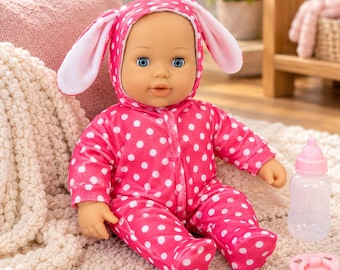 May include: A baby doll with blue eyes wearing a bright pink hooded onesie featuring white polka dots and long bunny ears attached to the hood. The doll is accompanied by a small, clear plastic baby bottle with a pink cap and a pink pacifier.