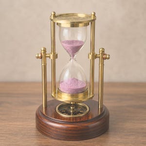 May include: A brass hourglass with purple sand held in a rotating frame supported by two vertical pillars. The base is a circular dark wood stand featuring an integrated magnetic compass with a black dial and gold-colored directional markings.